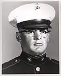 Head of a heavyset young white man with wire-frame glasses wearing a white peaked cap with black visor and a dark military jacket.