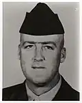 Head of a white man wearing a garrison cap and a dark military jacket over a light-colored shirt.