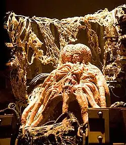 Ярко освещённое осьминогоподобное существо в окружении различного оборудования, покрытого волокнистым органическим материалом, смотрит вверх своим единственным глазом.