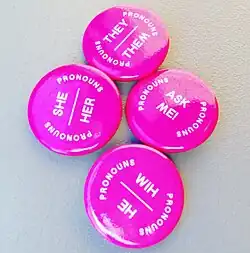 Four pin-on metal badges with labels «He Him», «She Her», «They Them», and «Ask Me!»