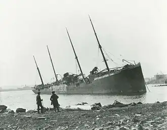 Two men observe a large beached ship with «Belgian Relief» painted on her side
