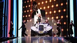 A group of performers on stage. Central to them, a blond female rides on a white car. She is flanked by dancers in black bondage style costumes. Behind them a background full of lights, is displayed.