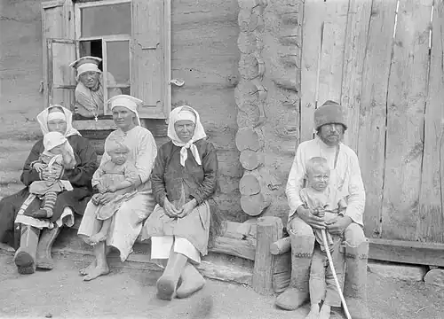 Пожилые и дети отдыхают у стены дома.