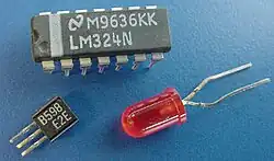 A small square plastic piece with three parallel wire protrusions on one side; a larger rectangular plastic chip with multiple plastic and metal pin-like legs; and a small red light globe with two long wires coming out of its base.