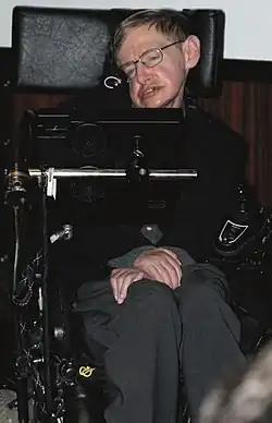A colored photograph of a man, sitting in an wheelchair with his arms folded. He is wearing glasses and is looking from left to right.